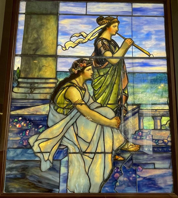 A girl and a boy on a terrace with balustrade, both in classical dress, the boy seated, the girl standing and playing a flute