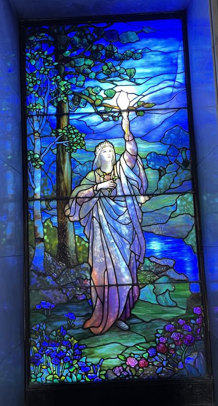 Seaver Memorial Window:  female figure holding aloft a blazing torch standing by creek in wooded area